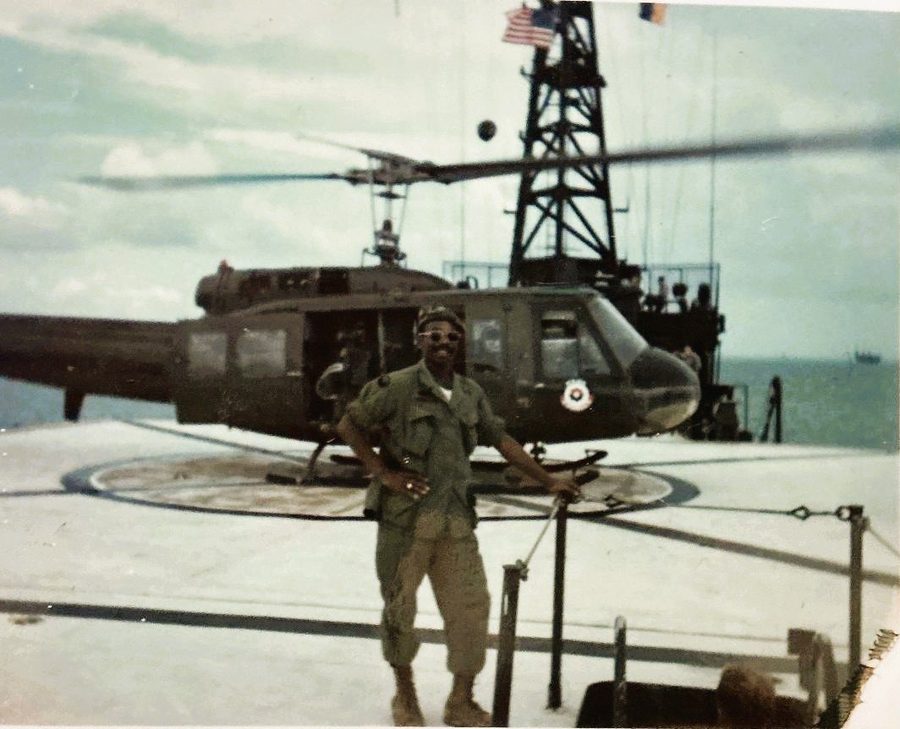 Leon Collier in fatigues and sunglasses standing before a UH-1 Huey helicopter on a ship's flight deck, an American flag flying behind him.