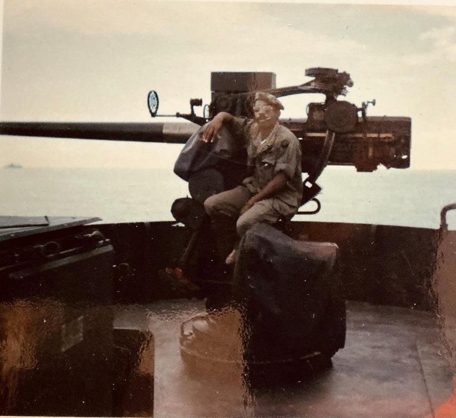 Leon Collier seated at a large deck gun aboard ship, the open sea behind him.