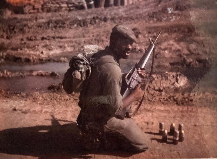 Leon Collier kneeling in the field in fatigues, holding his rifle, a rucksack on his back.