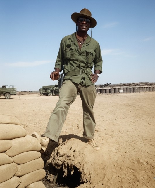 Leon Collier standing outdoors in fatigues and a wide-brim bush hat, one boot resting on a sandbagged bunker, military trucks and a desert base behind him.