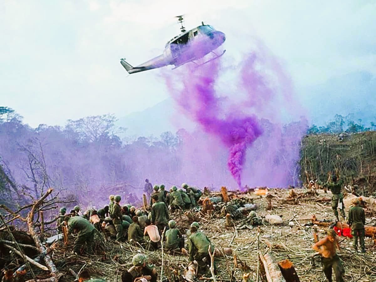 A UH-1 Huey helicopter flying overhead trailing purple signal smoke above American soldiers gathered on a cleared hillside.